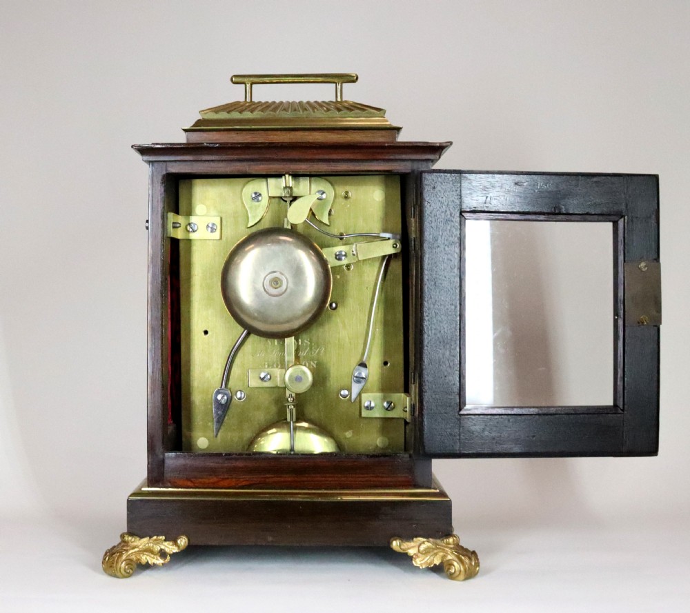 A mahogany and brass bracket clock featuring a white enamel dial with Roman numerals, gilt brass corner mounts, and scroll feet. View VII