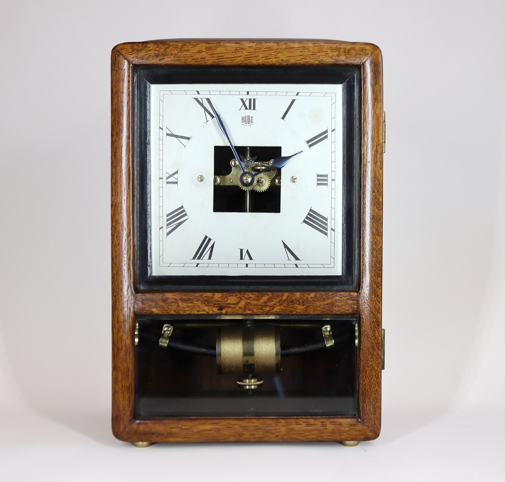 An Art Deco oak-cased mantel clock featuring a square white dial with Roman numerals and a clear glass window revealing the brass pendulum mechanism below.