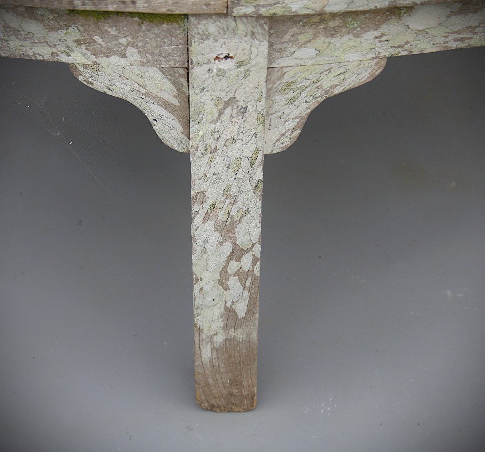 A curved wooden slatted garden bench with weathered grey finish and natural moss growth, designed to encircle a tree trunk. View III