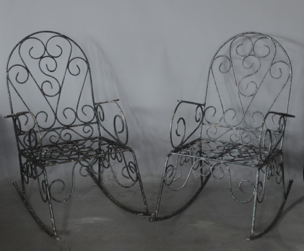 A pair of dark wrought iron garden rocking chairs featuring elaborate scrollwork patterns on the backrests, armrests, and bases.