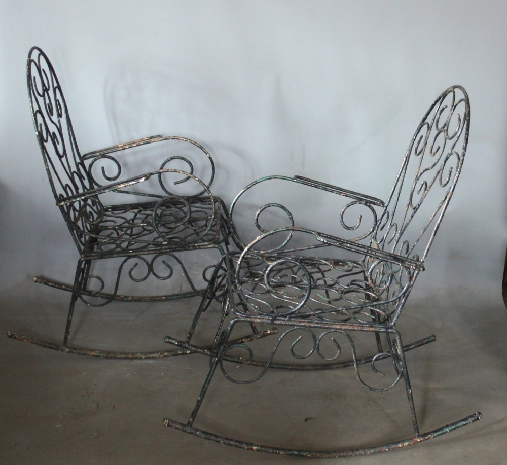 A pair of dark wrought iron garden rocking chairs featuring elaborate scrollwork patterns on the backrests, armrests, and bases. View II