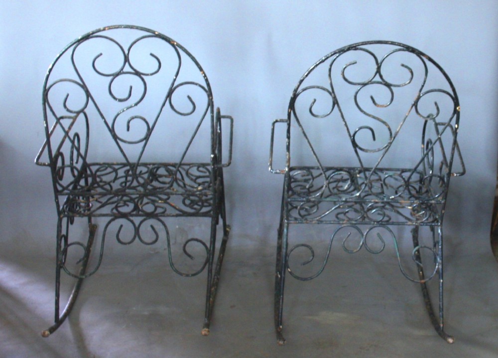 A pair of dark wrought iron garden rocking chairs featuring elaborate scrollwork patterns on the backrests, armrests, and bases. View III