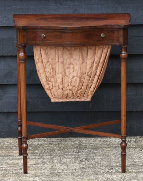fig 1. An Edwardian mahogany sewing table featuring a serpentine front with line inlay, slender turned legs connected by an X-frame stretcher, and a pleated cloth basket suspended underneath.