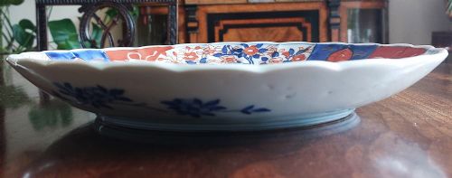 fig 1. A circular Japanese Imari porcelain plate featuring a scalloped edge, decorated in panelled cobalt blue, iron red, and floral motifs surrounding a central vase illustration.