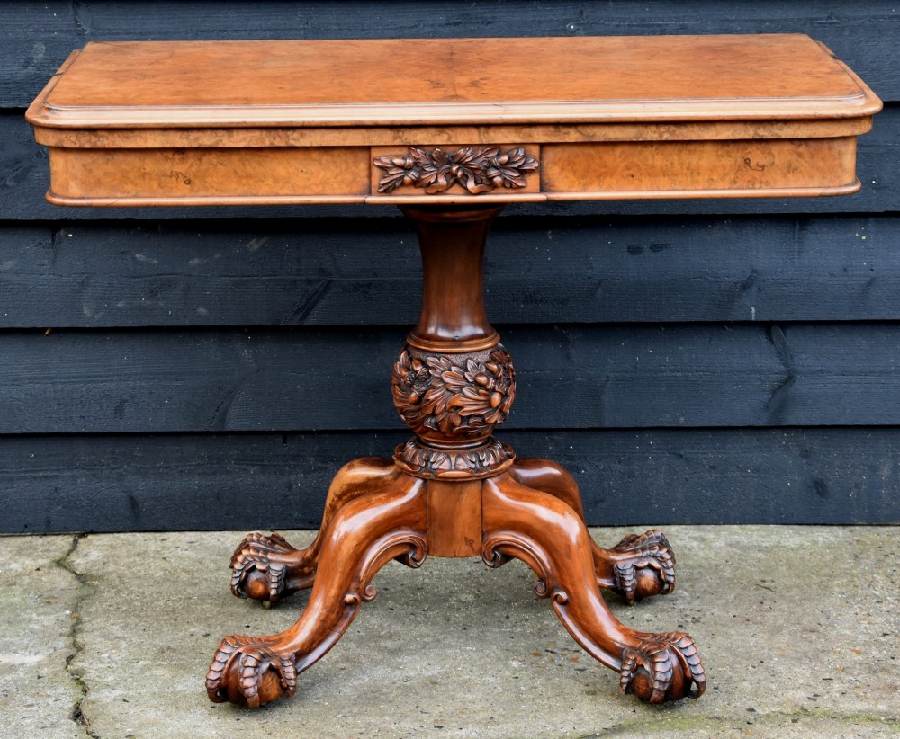 A Victorian burr walnut foldover card table featuring a rectangular swivel top, carved apron accent, turned pedestal stem with floral carving, and four cabriole legs ending in claw and ball feet.