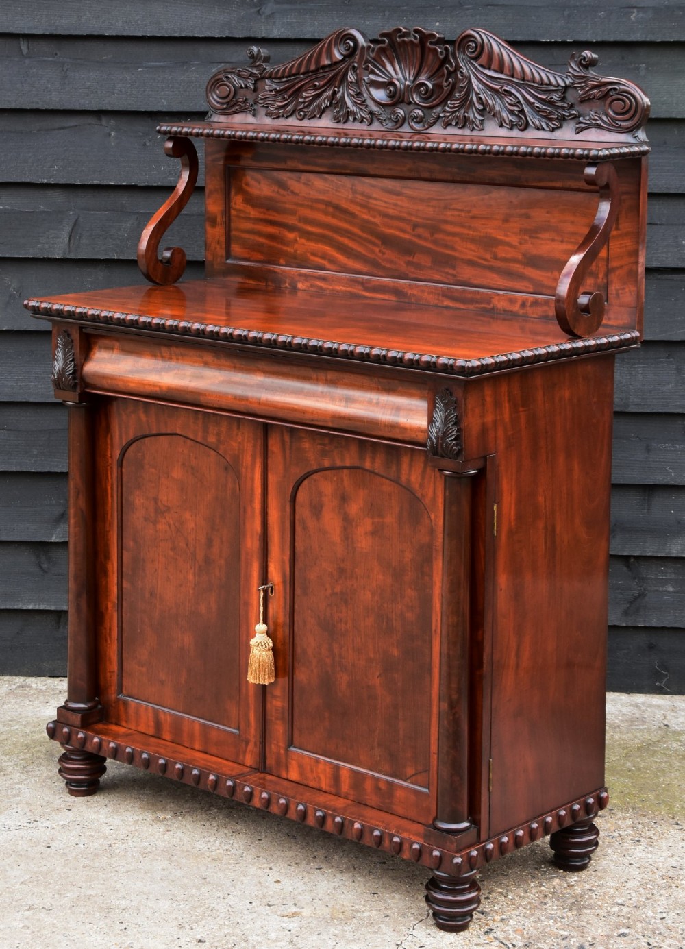 A Regency mahogany chiffonier side cabinet featuring a carved cresting gallery shelf, flame mahogany cupboard doors, and turned feet.