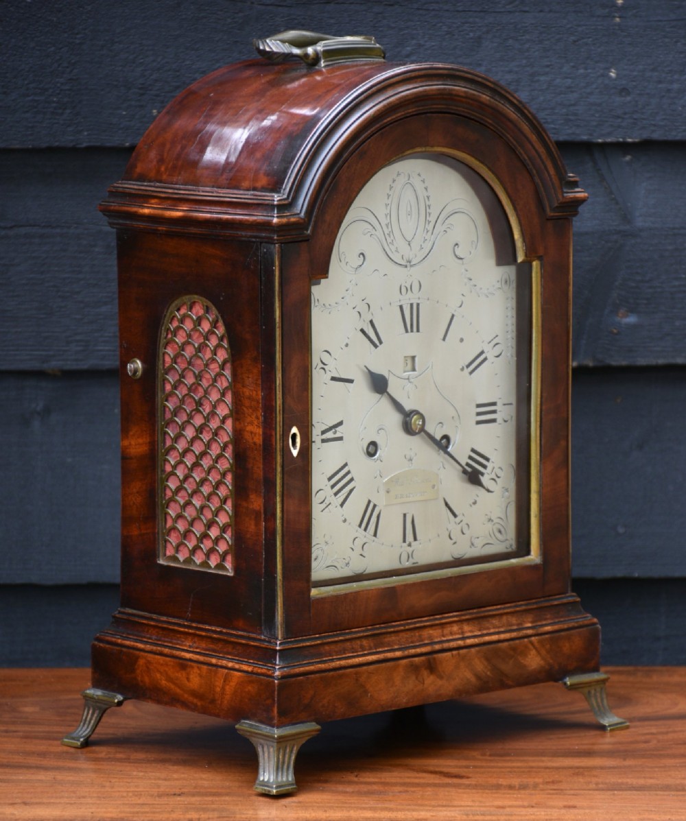 A Georgian mahogany bracket clock featuring an arched top case, top brass carrying handle, pierced side fretwork, engraved dial with Roman numerals, and brass bracket feet.