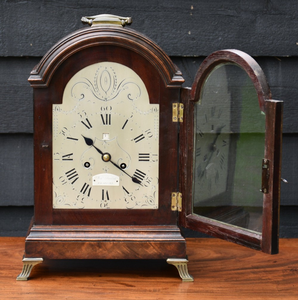 A Georgian mahogany bracket clock featuring an arched top case, top brass carrying handle, pierced side fretwork, engraved dial with Roman numerals, and brass bracket feet. View II
