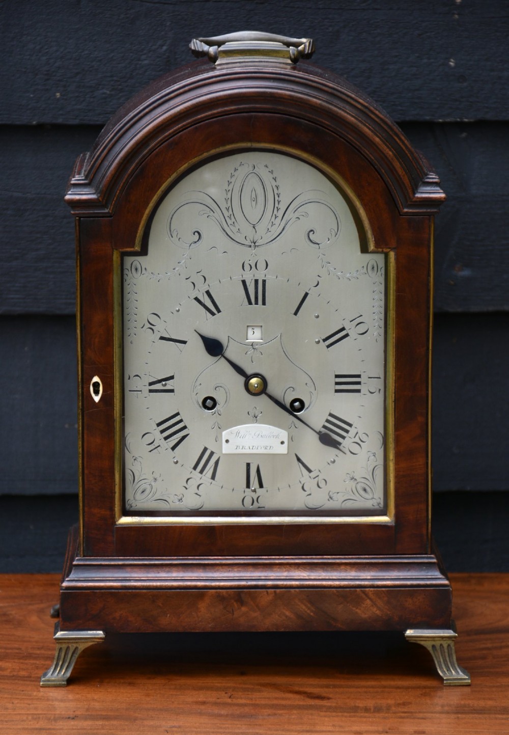 A Georgian mahogany bracket clock featuring an arched top case, top brass carrying handle, pierced side fretwork, engraved dial with Roman numerals, and brass bracket feet. View III