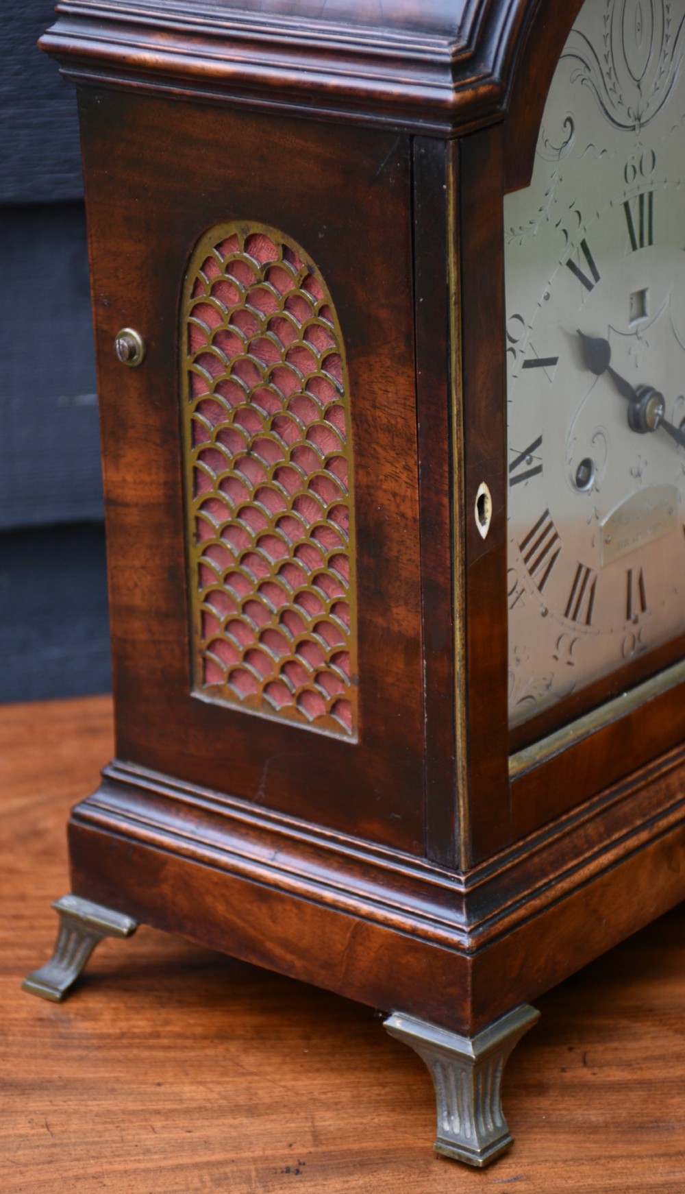 A Georgian mahogany bracket clock featuring an arched top case, top brass carrying handle, pierced side fretwork, engraved dial with Roman numerals, and brass bracket feet. View V