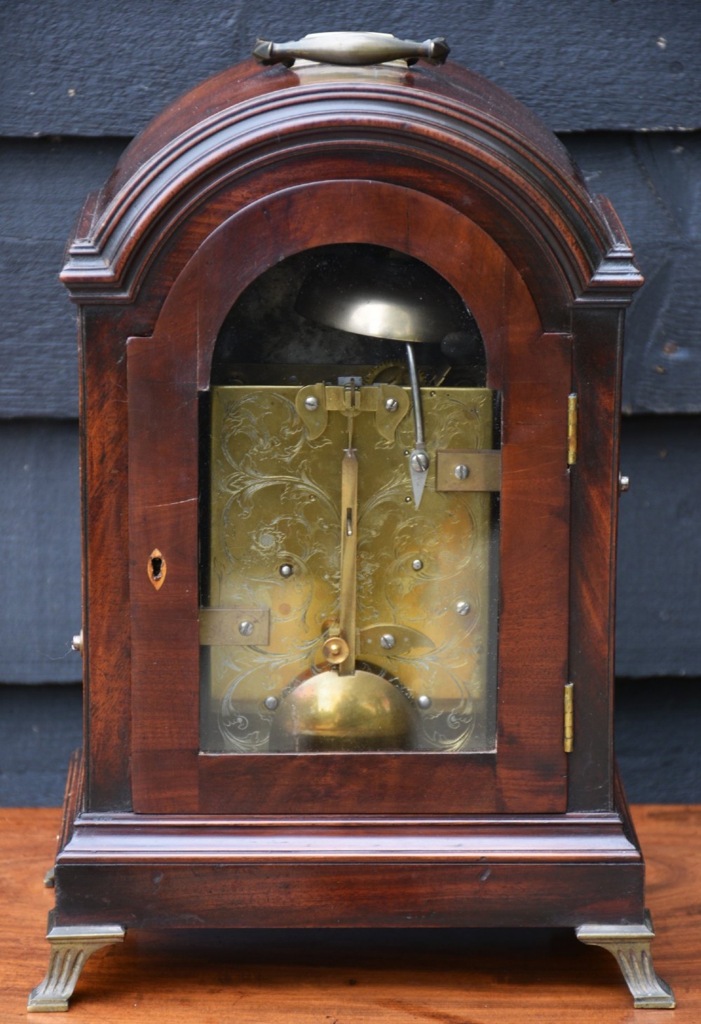 A Georgian mahogany bracket clock featuring an arched top case, top brass carrying handle, pierced side fretwork, engraved dial with Roman numerals, and brass bracket feet. View VI