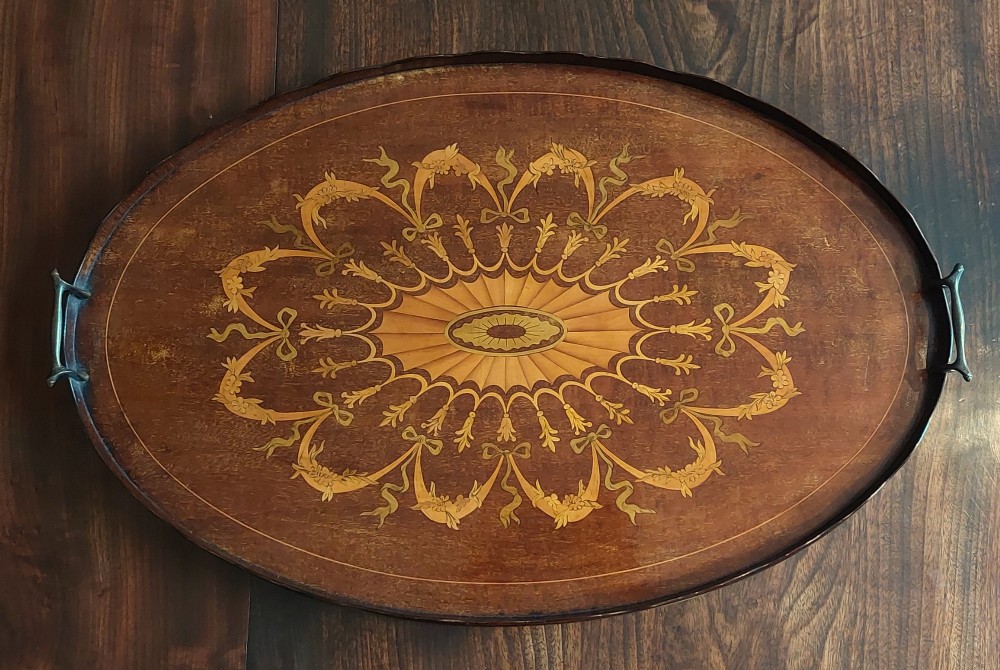 An oval mahogany serving tray featuring intricate central floral marquetry inlay, ribbon swags, and two cast brass side handles.