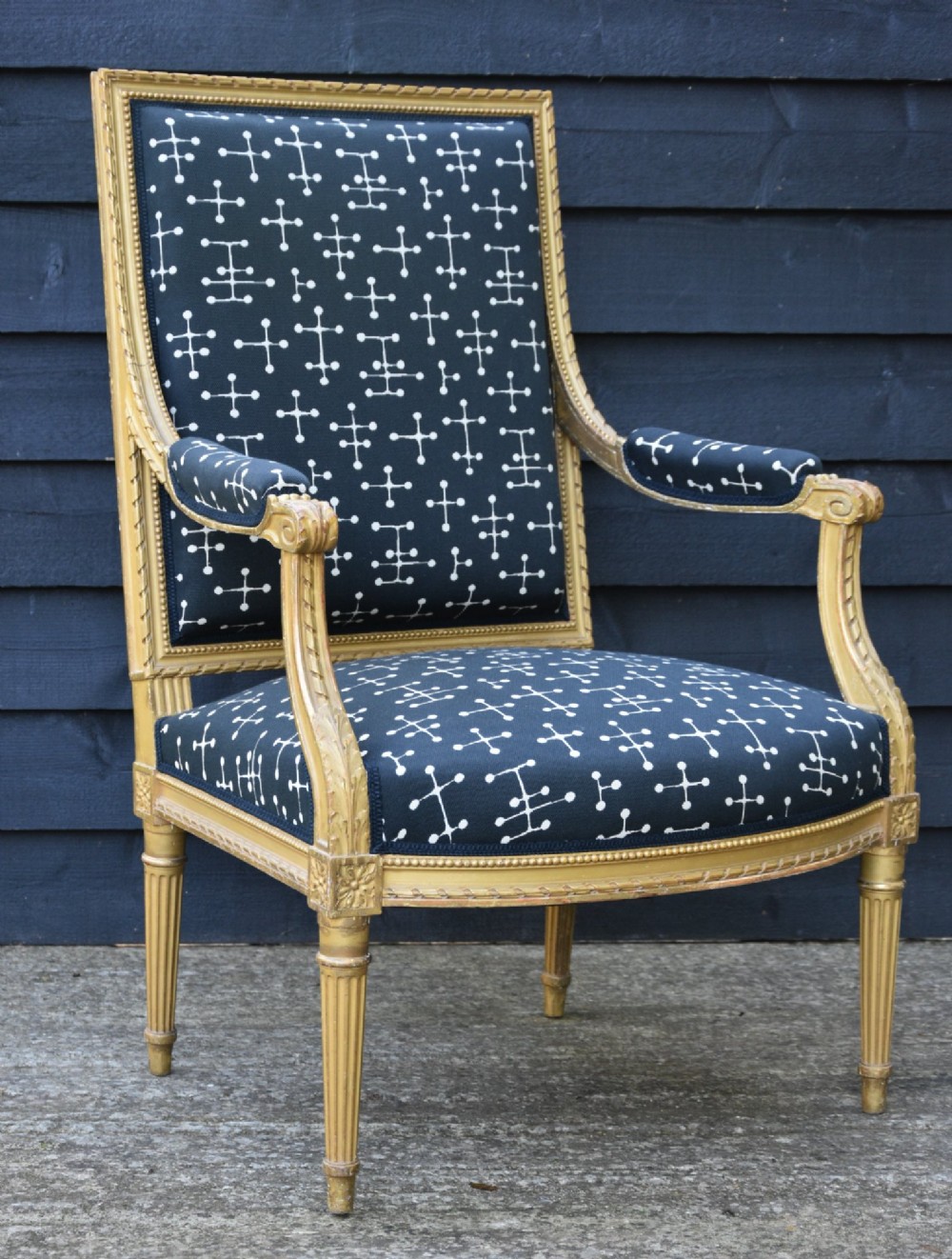 A French giltwood open armchair featuring a rectangular padded backrest, carved fluted legs, and navy blue upholstery with white geometric patterns.