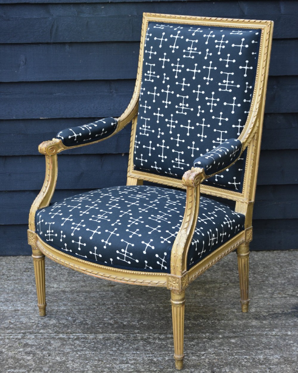 A French giltwood open armchair featuring a rectangular padded backrest, carved fluted legs, and navy blue upholstery with white geometric patterns. View IV