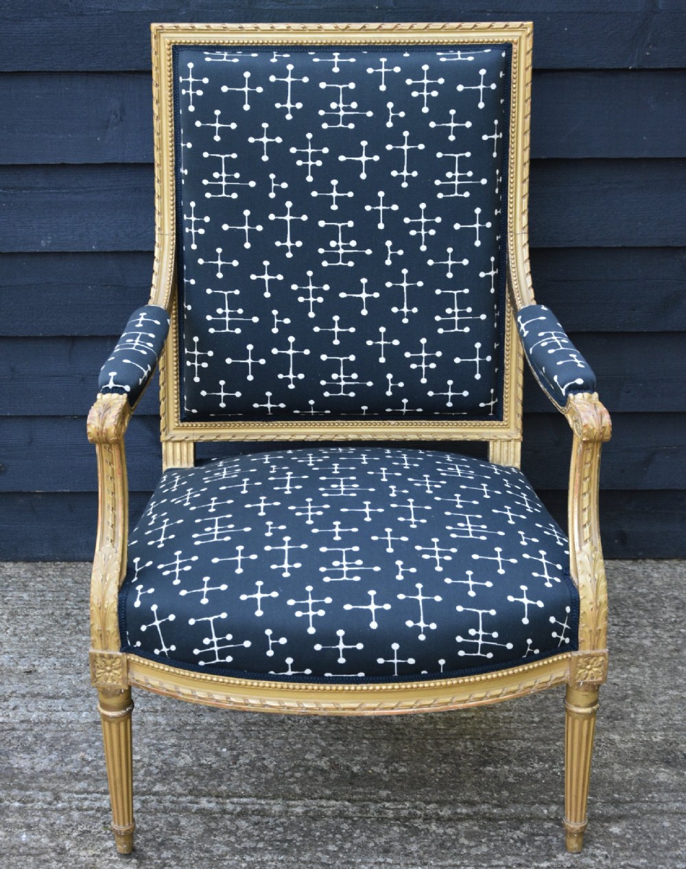 A French giltwood open armchair featuring a rectangular padded backrest, carved fluted legs, and navy blue upholstery with white geometric patterns. View V