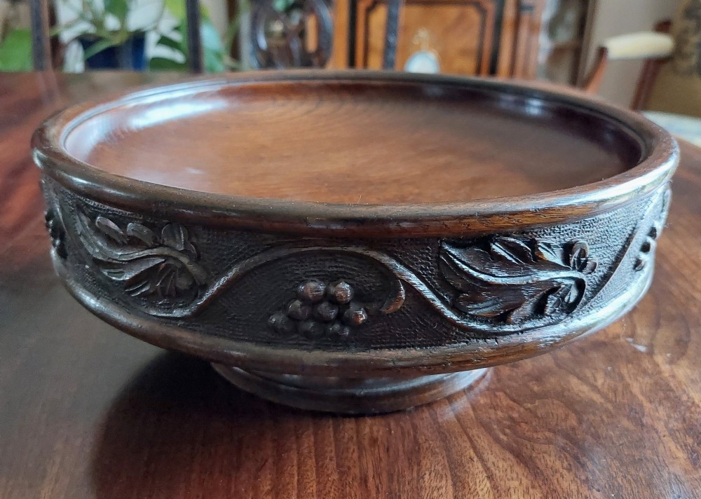 A circular dark carved oak fruit bowl on a low pedestal base decorated with a relief band of carved grapevines. View II