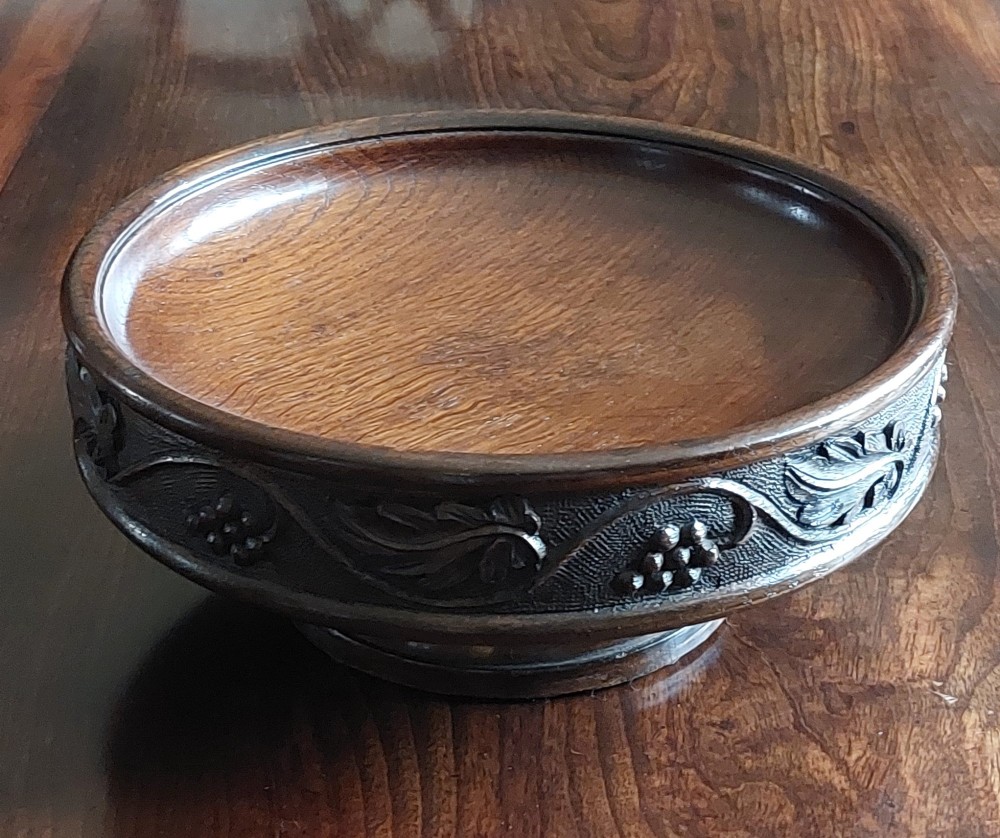 A circular dark carved oak fruit bowl on a low pedestal base decorated with a relief band of carved grapevines. View V