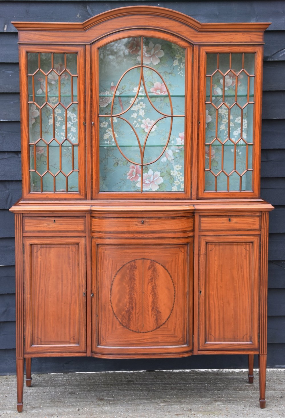 An Edwardian satinwood display cabinet featuring three astragal glazed doors, floral fabric interior lining, and inlaid lower cupboard doors raised on square tapered legs.