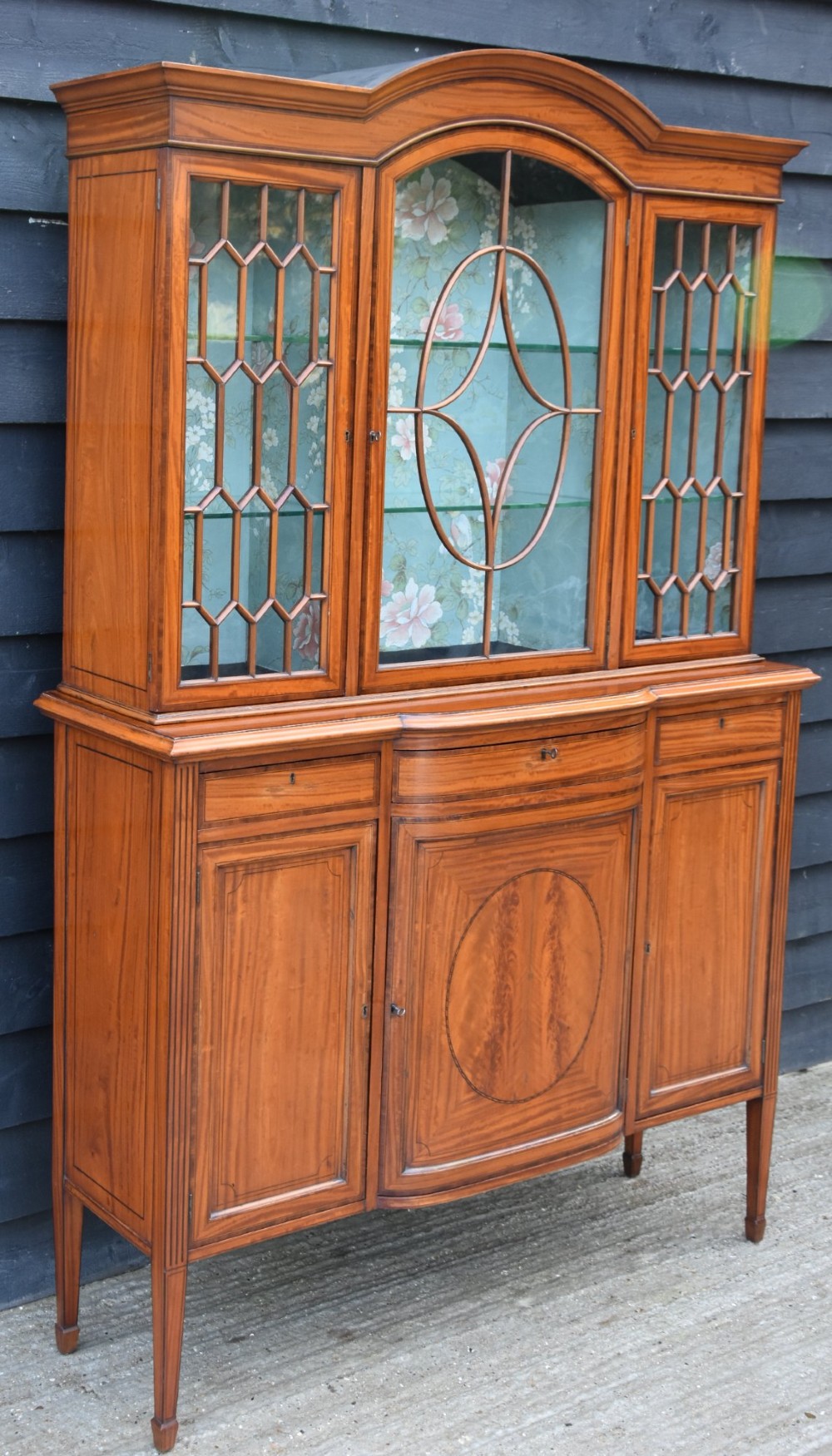 An Edwardian satinwood display cabinet featuring three astragal glazed doors, floral fabric interior lining, and inlaid lower cupboard doors raised on square tapered legs. View IV
