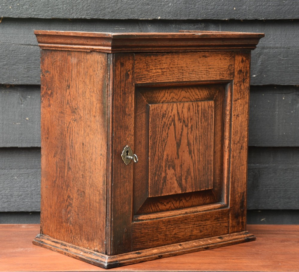 An early 18th-century oak spice cupboard featuring a single fielded panel door, brass escutcheon, and moulded top and base trim. View VII