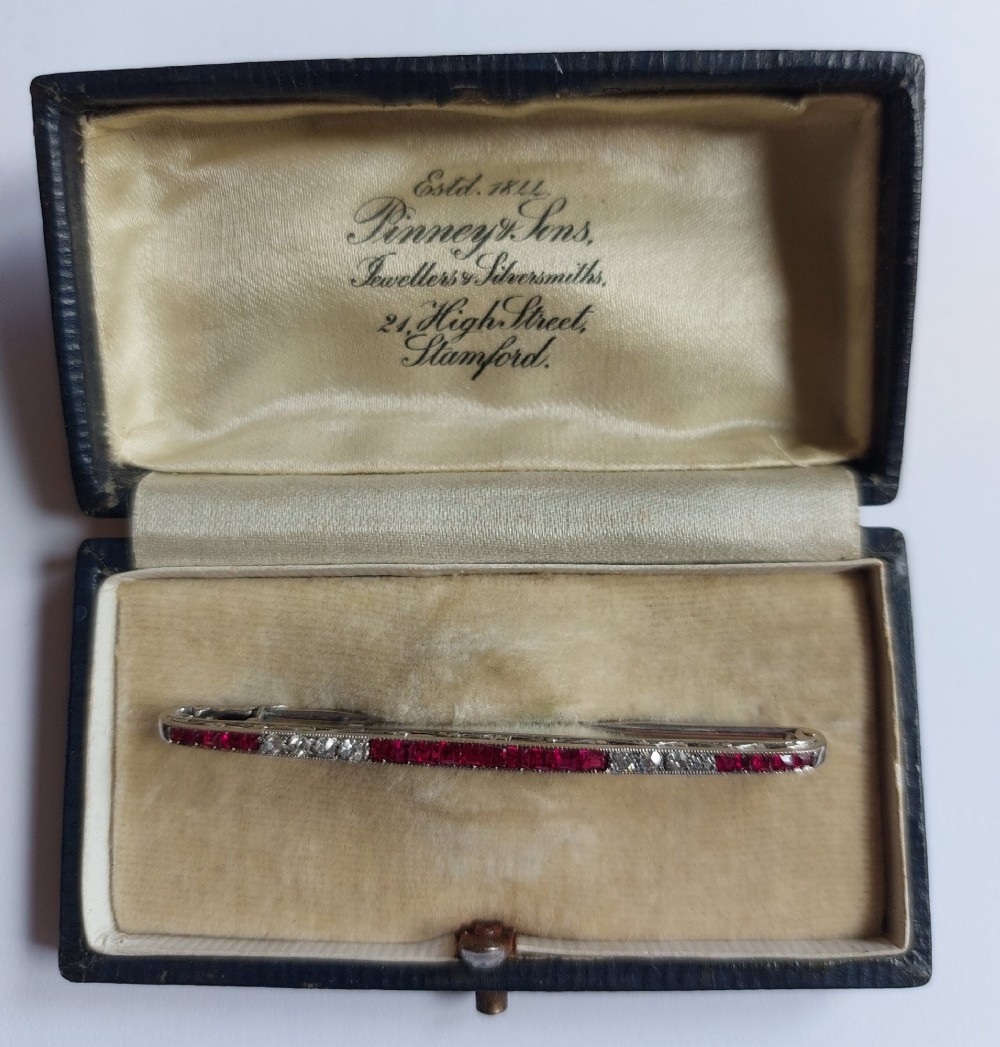 An Art Deco slender bar brooch crafted in white metal, channel-set with square red rubies interspersed with small round diamonds with millegrain detailing. View VII