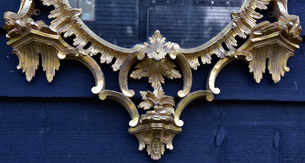 An 18th-century Georgian giltwood wall mirror featuring an oval mirror plate surrounded by an intricately carved gold leaf frame with acanthus leaf cresting and C-scroll motifs. View V
