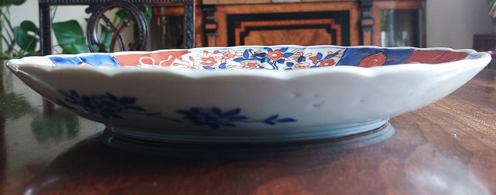 A circular Japanese Imari porcelain plate featuring a scalloped rim, hand-painted cobalt blue and iron-red floral motifs, and broad panels arranged around a central vase design. View III