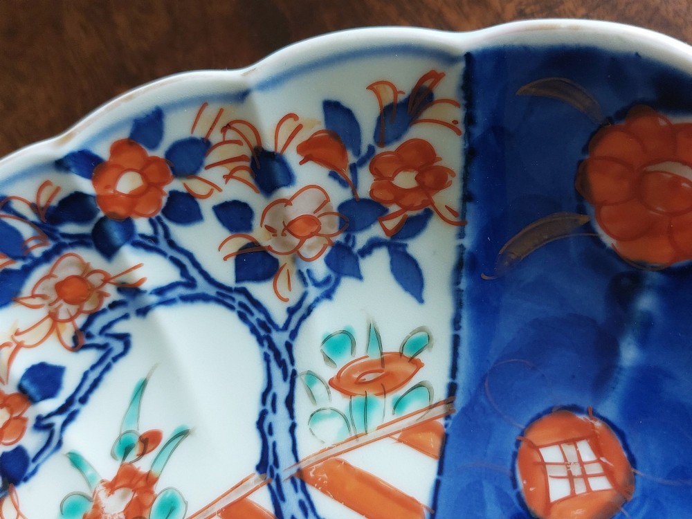 A circular Japanese Imari porcelain plate featuring a scalloped rim, hand-painted cobalt blue and iron-red floral motifs, and broad panels arranged around a central vase design. View VI