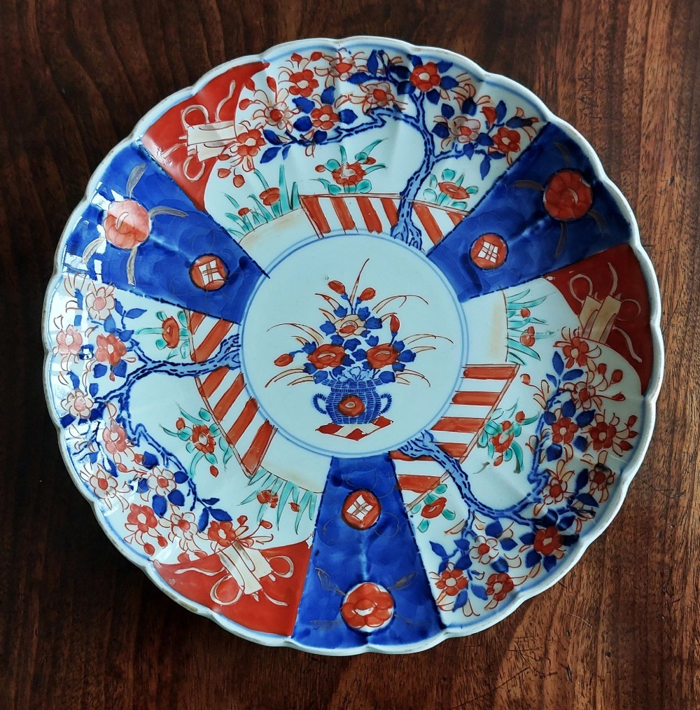 A circular Japanese Imari porcelain plate featuring a scalloped rim, hand-painted cobalt blue and iron-red floral motifs, and broad panels arranged around a central vase design.