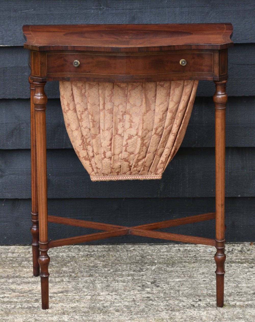 An Edwardian mahogany sewing table featuring a serpentine front with line inlay, slender turned legs connected by an X-frame stretcher, and a pleated cloth basket suspended underneath. View II