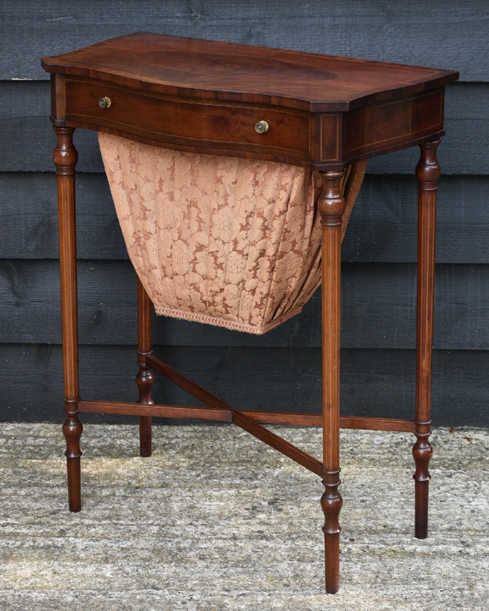 An Edwardian mahogany sewing table featuring a serpentine front with line inlay, slender turned legs connected by an X-frame stretcher, and a pleated cloth basket suspended underneath. View V