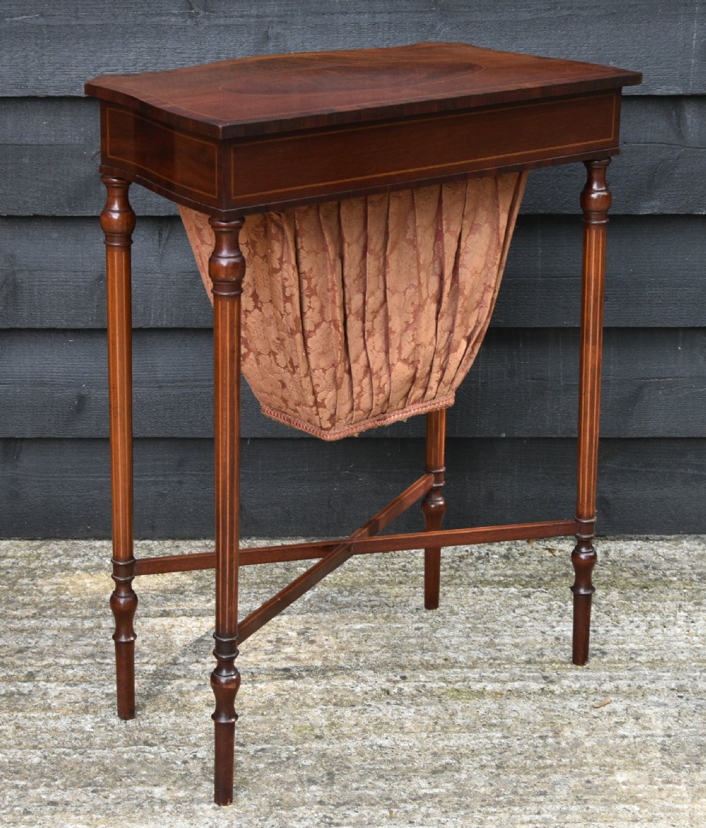 An Edwardian mahogany sewing table featuring a serpentine front with line inlay, slender turned legs connected by an X-frame stretcher, and a pleated cloth basket suspended underneath. View VI
