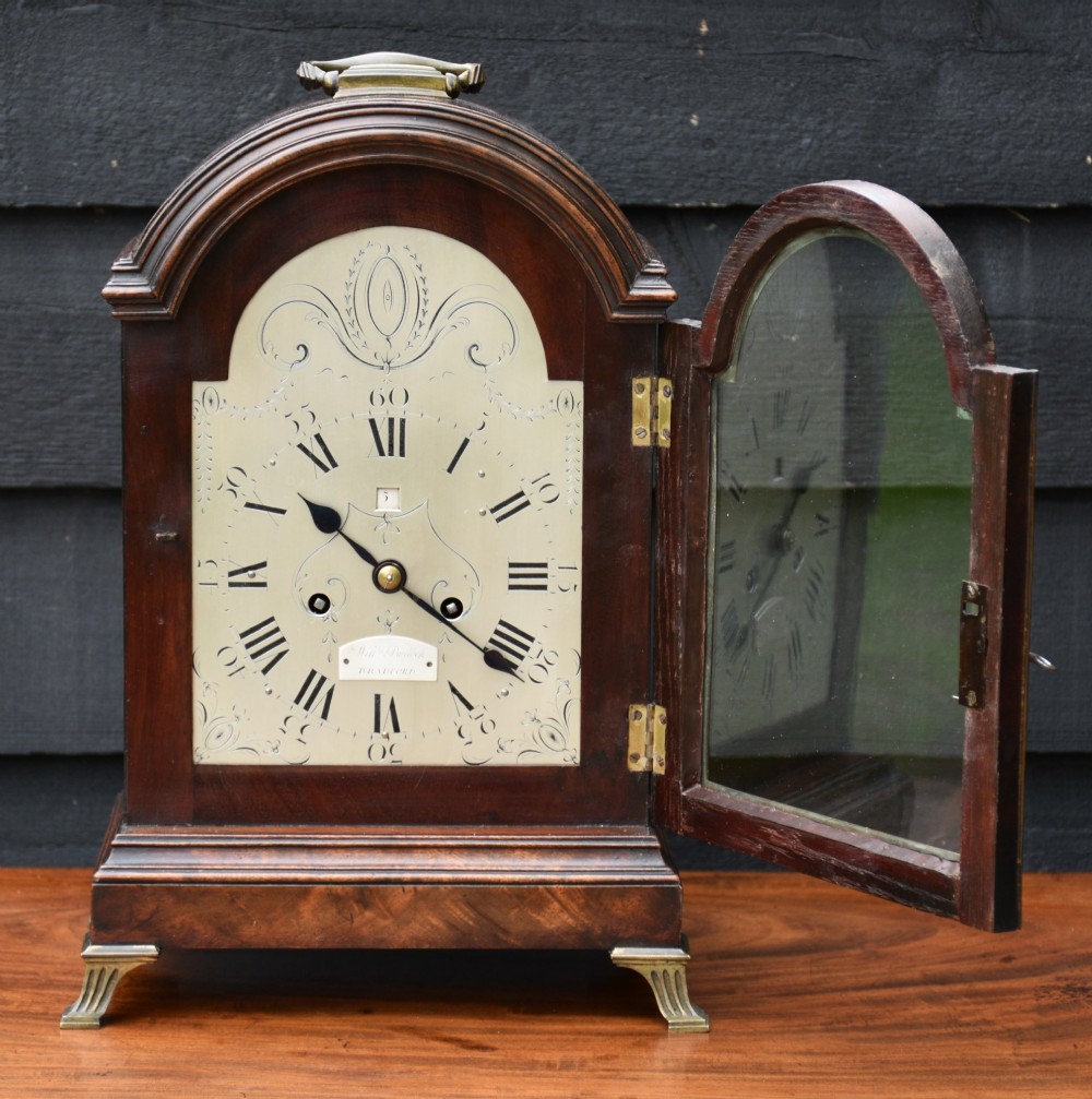 An antique Georgian mahogany bracket clock featuring an arched top case, brass carrying handle, engraved silvered dial face, and fluted brass feet. View II