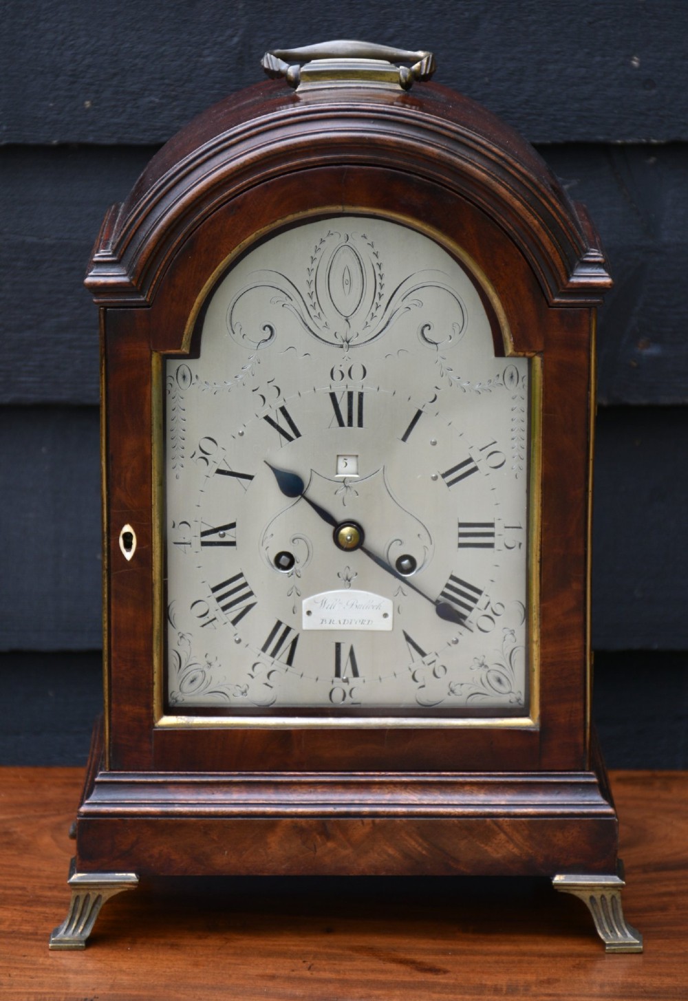 An antique Georgian mahogany bracket clock featuring an arched top case, brass carrying handle, engraved silvered dial face, and fluted brass feet. View VIII