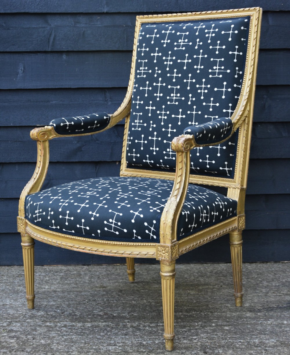 A French Louis XVI style giltwood open armchair upholstered in dark blue fabric with white geometric motifs, featuring fluted legs and carved leaf moulding. View IV