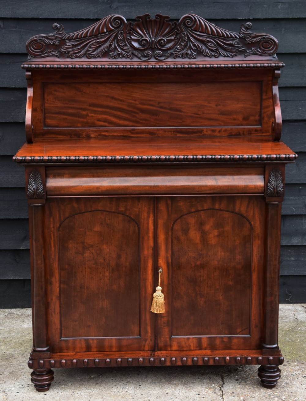 A Regency mahogany chiffonier cabinet featuring an ornate carved shell crest shelf, frieze drawer, two cupboard doors with flame mahogany panels, and turned feet.