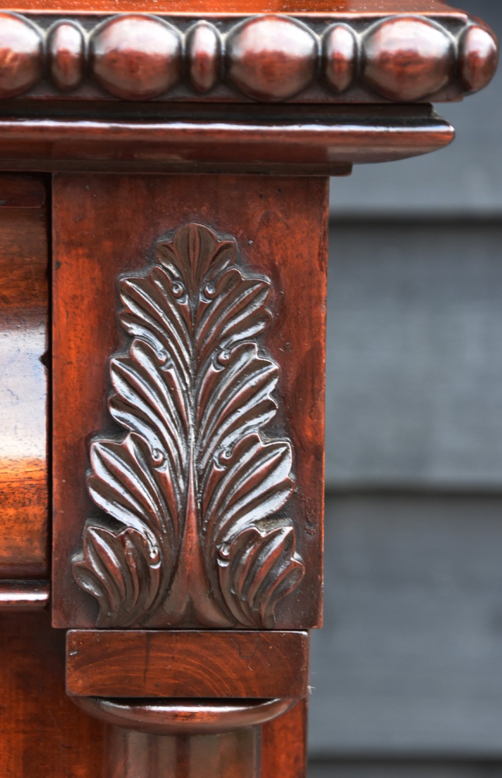 A Regency mahogany chiffonier cabinet featuring an ornate carved shell crest shelf, frieze drawer, two cupboard doors with flame mahogany panels, and turned feet. View III