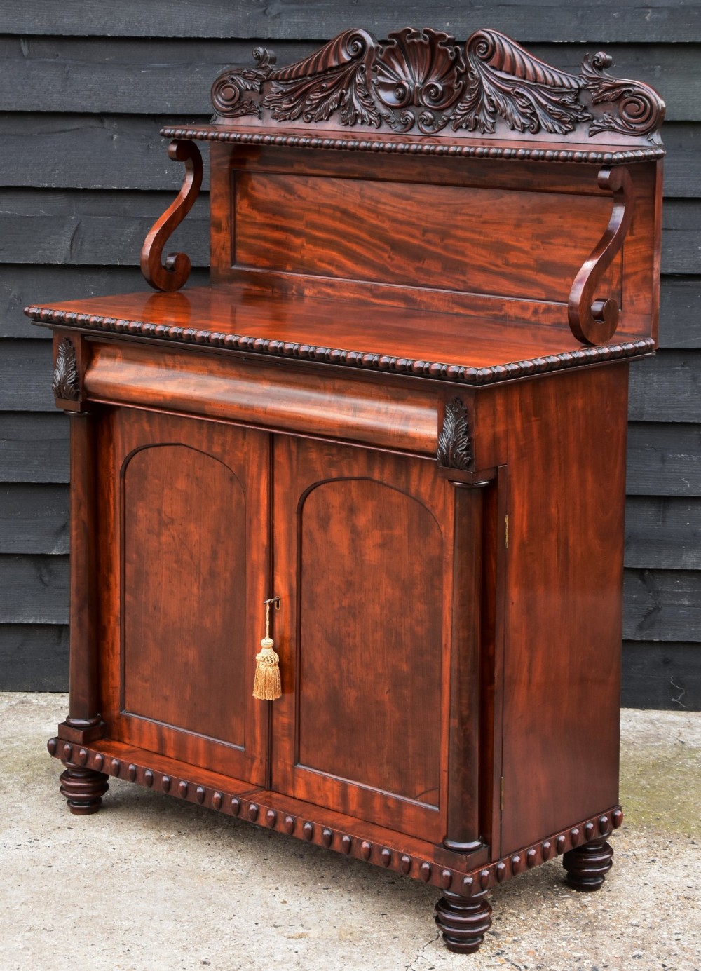 A Regency mahogany chiffonier cabinet featuring an ornate carved shell crest shelf, frieze drawer, two cupboard doors with flame mahogany panels, and turned feet. View V