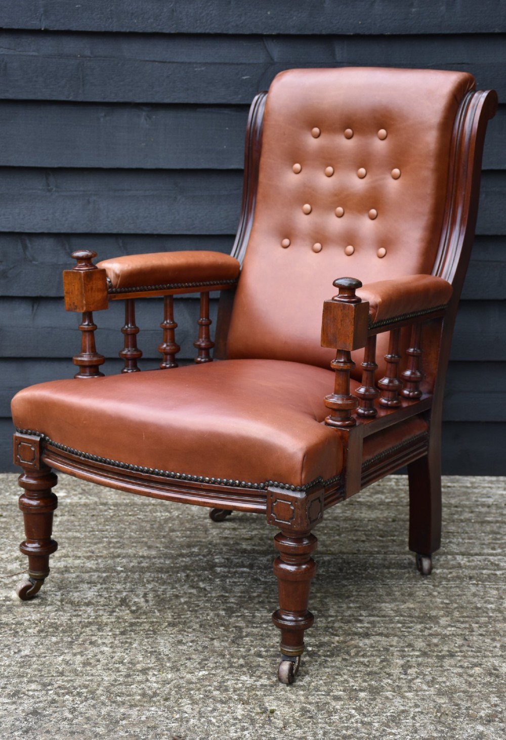 A Victorian mahogany library armchair featuring tan leather upholstery with button-tufting on the backrest, turned spindle arm supports, brass stud detailing, and turned front legs on castors. View VII