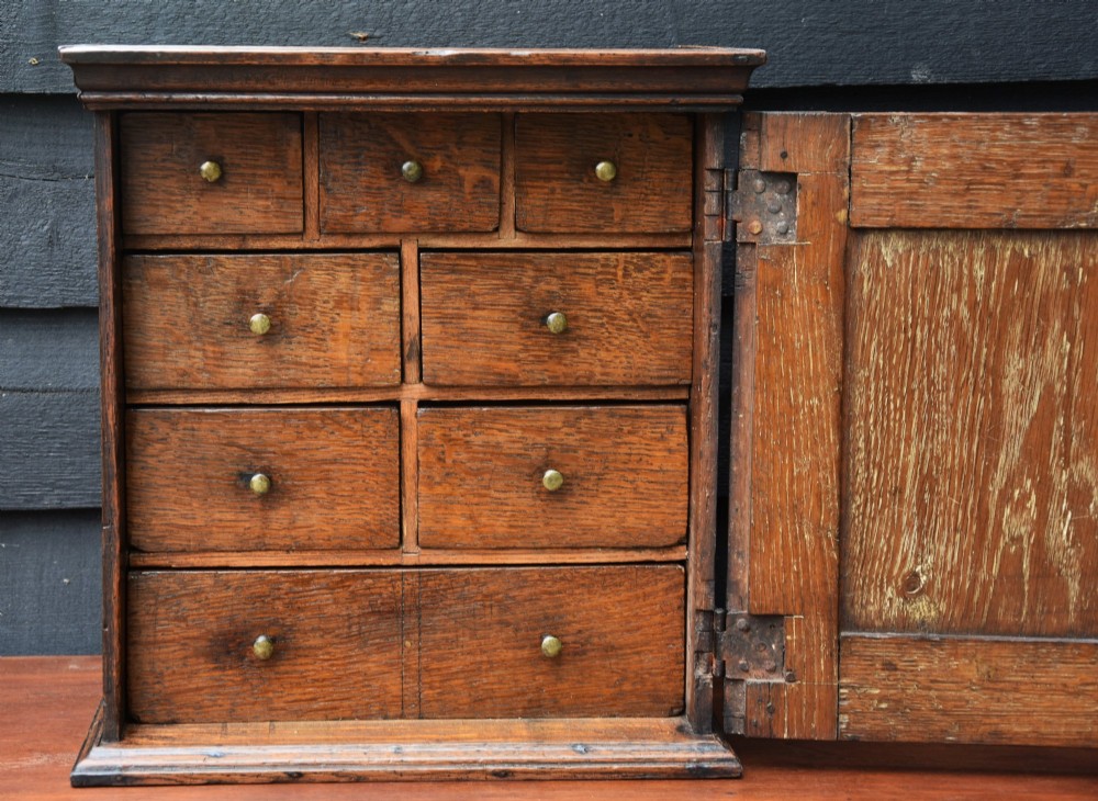 An early eighteenth-century oak spice cupboard featuring a single fielded panel door with a brass escutcheon and key, set on a moulded base. View XI