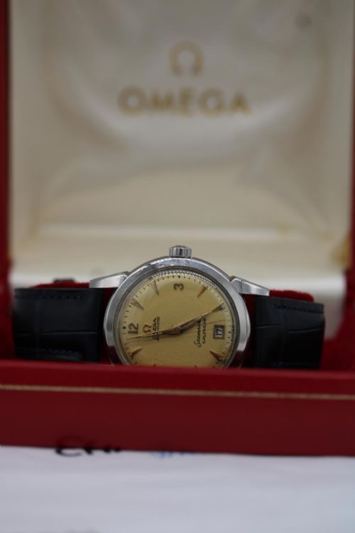 fig 7. A vintage 1952 Omega Seamaster Calendar wristwatch featuring a stainless steel case, textured gold-coloured dial with date display at six oclock, and a black leather strap.