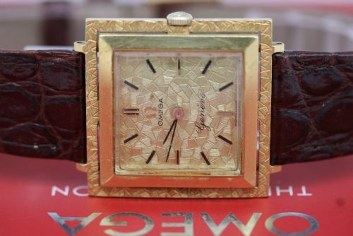 fig 1. A 1960s Omega Geneve manual wind wristwatch featuring an 18ct yellow gold cushion-shaped case, a dark brown dial with gold Roman numerals at twelve and six, and a brown leather strap.