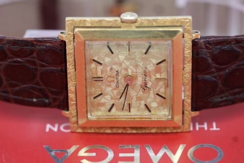 fig 6. A 1960s Omega Geneve manual wind wristwatch featuring an 18ct yellow gold cushion-shaped case, a dark brown dial with gold Roman numerals at twelve and six, and a brown leather strap.