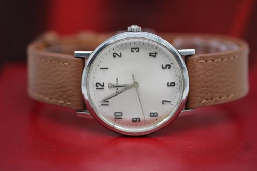 fig 7. A 1967 stainless steel Omega wristwatch featuring a white dial with black Arabic numerals and a tan brown leather strap.