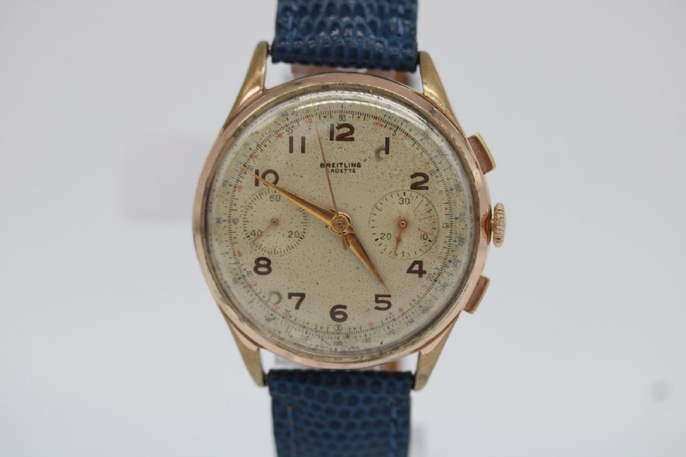 A 1950s Breitling Cadette chronograph wristwatch featuring a circular gold-plated case, twin sub-dials, Arabic numerals, and a blue textured leather strap.