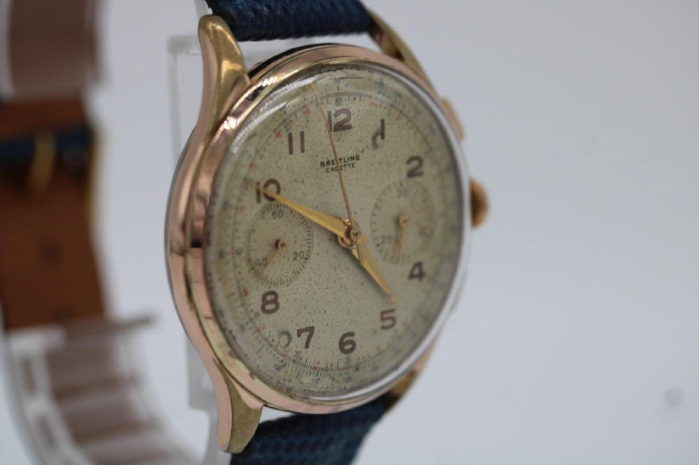 A 1950s Breitling Cadette chronograph wristwatch featuring a circular gold-plated case, twin sub-dials, Arabic numerals, and a blue textured leather strap. View VII