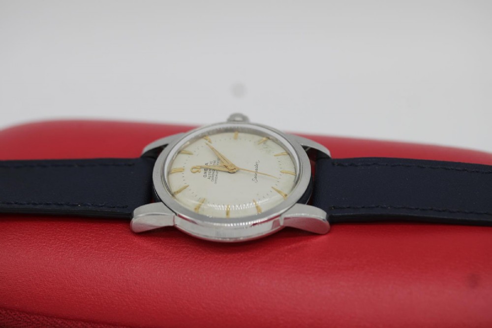 A vintage 1956 Omega Seamaster automatic wristwatch featuring a silver-coloured stainless steel case, off-white dial with gold-tone markers, and a navy blue leather strap. View II