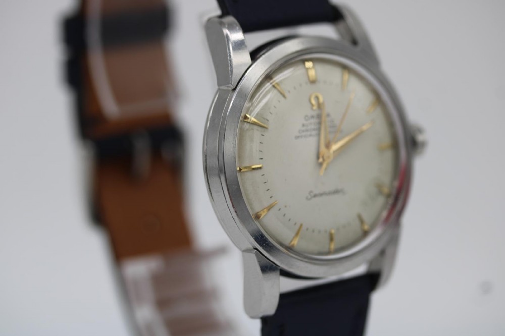 A vintage 1956 Omega Seamaster automatic wristwatch featuring a silver-coloured stainless steel case, off-white dial with gold-tone markers, and a navy blue leather strap. View III