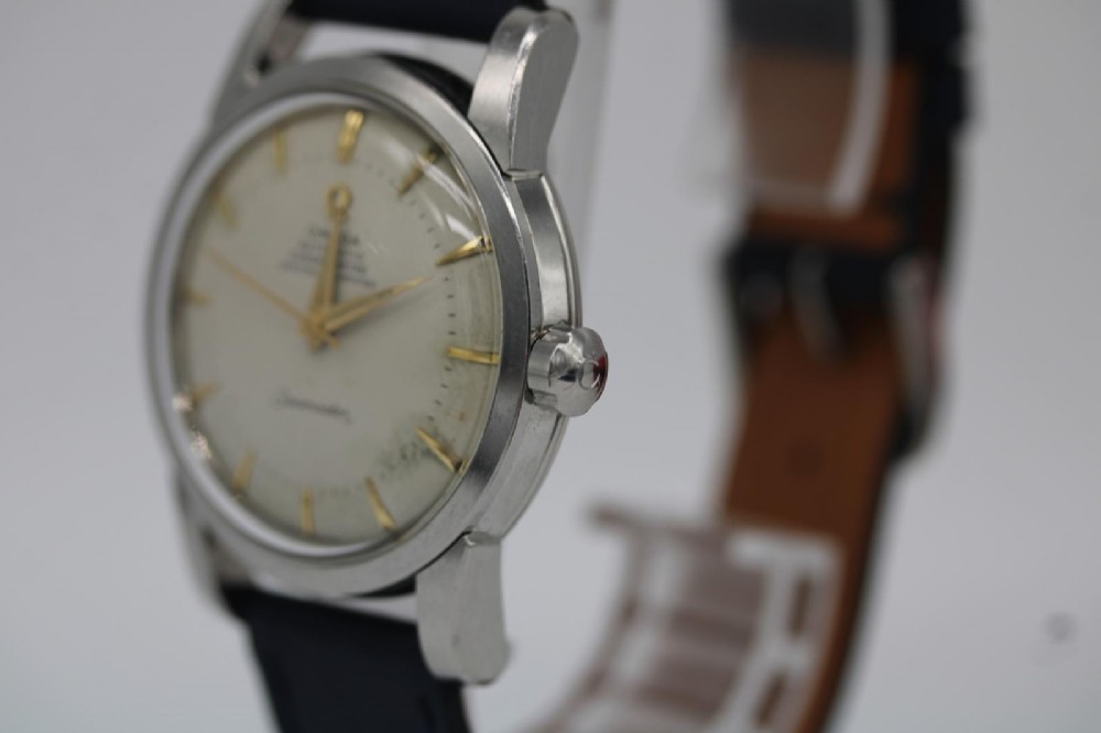 A vintage 1956 Omega Seamaster automatic wristwatch featuring a silver-coloured stainless steel case, off-white dial with gold-tone markers, and a navy blue leather strap. View VII