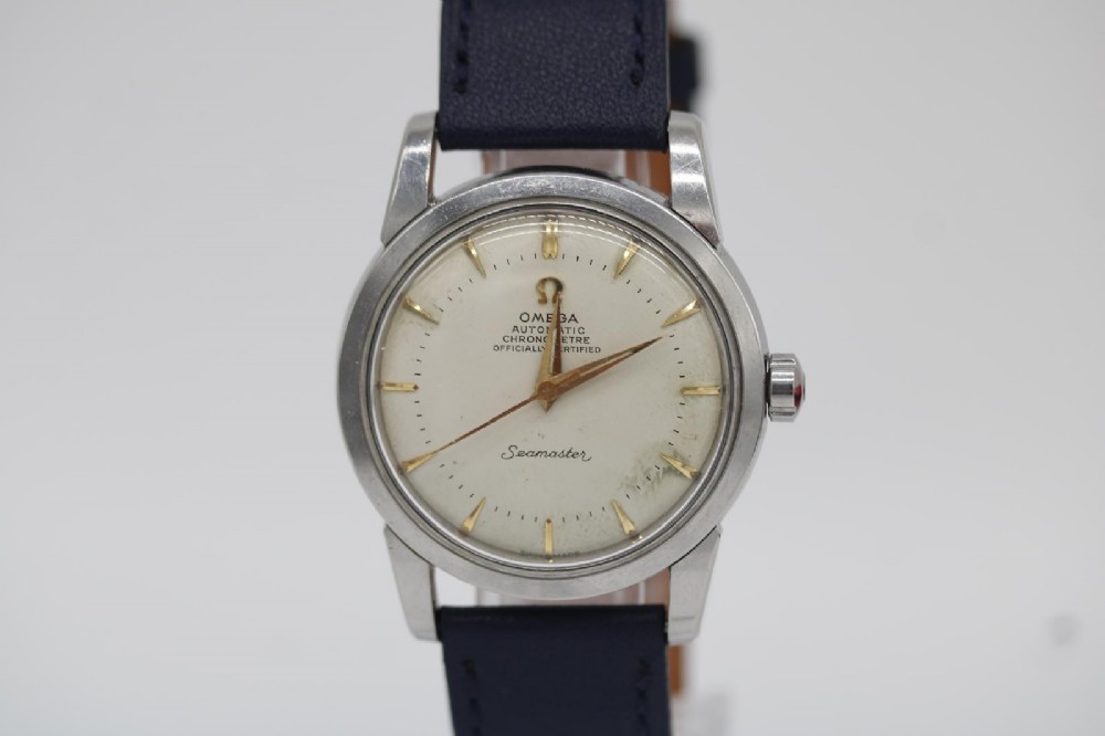 A vintage 1956 Omega Seamaster automatic wristwatch featuring a silver-coloured stainless steel case, off-white dial with gold-tone markers, and a navy blue leather strap.